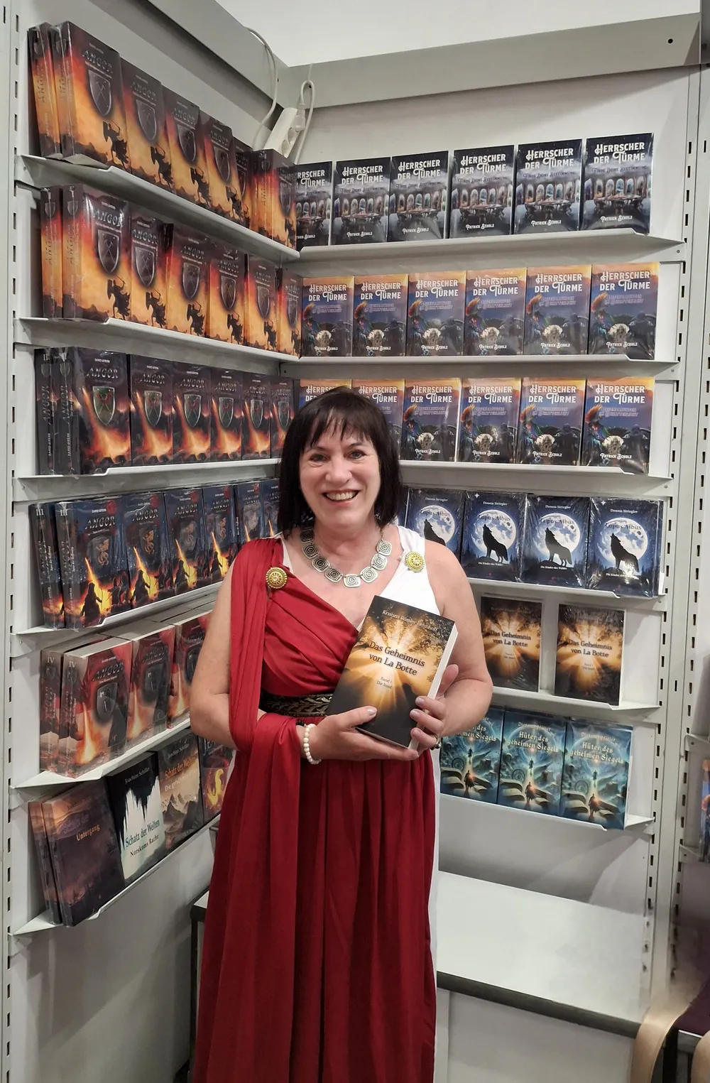 Kristina Rössler-Lehnhoff in rotem Gewand auf der Frankfurter Buchmesse, vor Bücherregalen, „Das Geheimnis von La Botte“ in der Hand